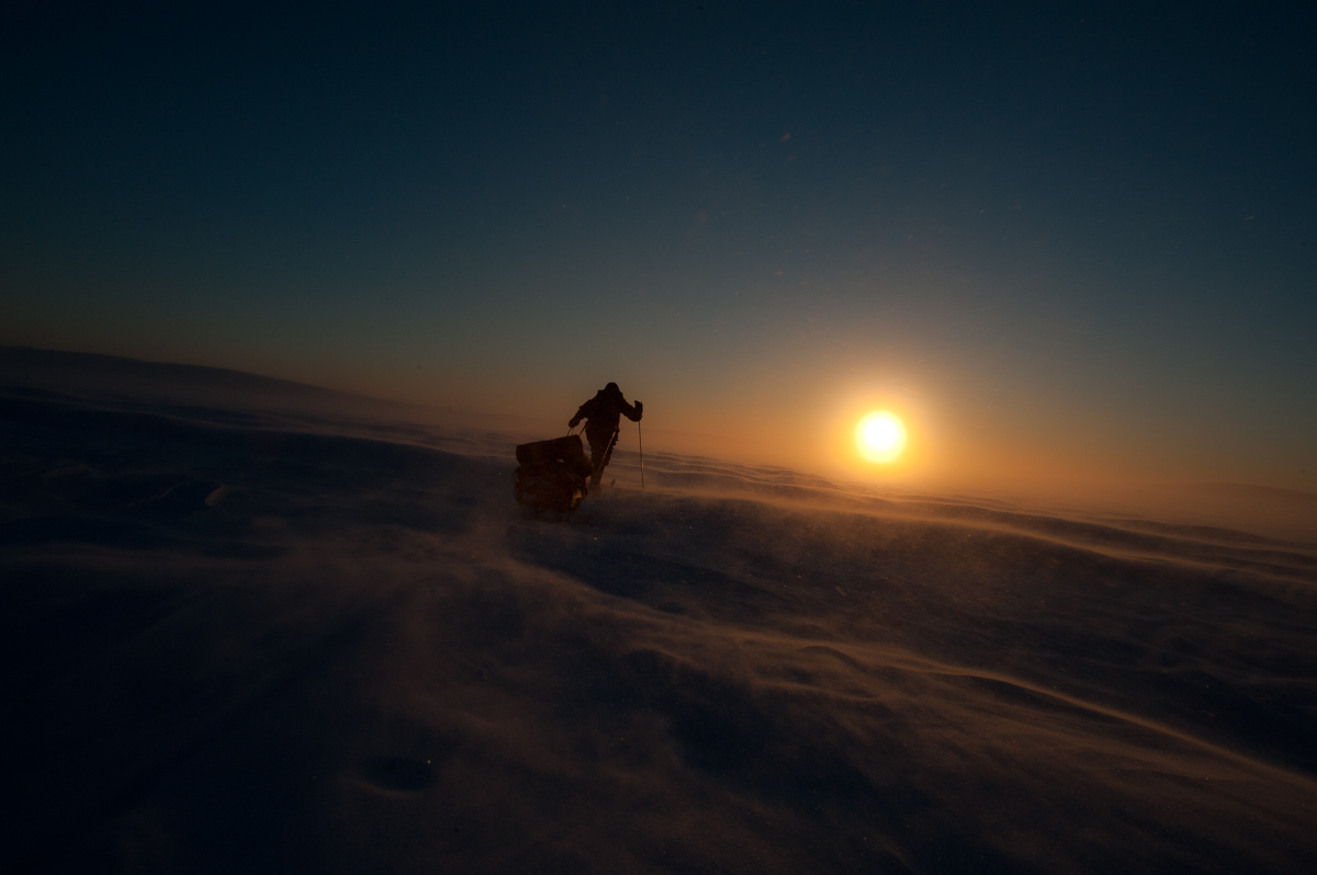 A skier pulling a sled into a snow drift at sunset