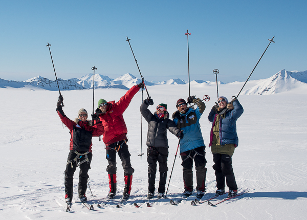 Five skiers in colourful jackets raising their poles on a sunny glacier